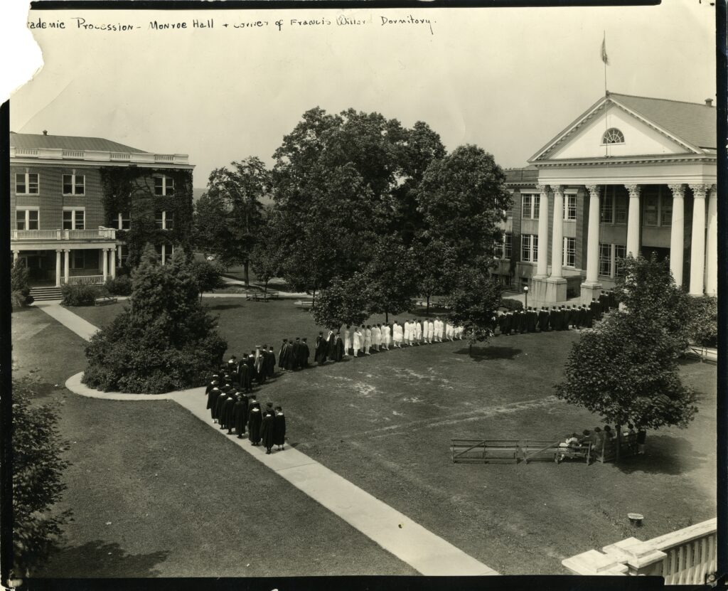 A black and white photo showing an academic procession walking into Monroe Hall. Presumably students and faculty are walking two by two in black and white robes. There is handwritten text at the top of the picture that says "Academic Procession - Monroe Hall + Corner of Francis Williard Dormitory." The photo is taken from a higher up floor of Virginia Hall.