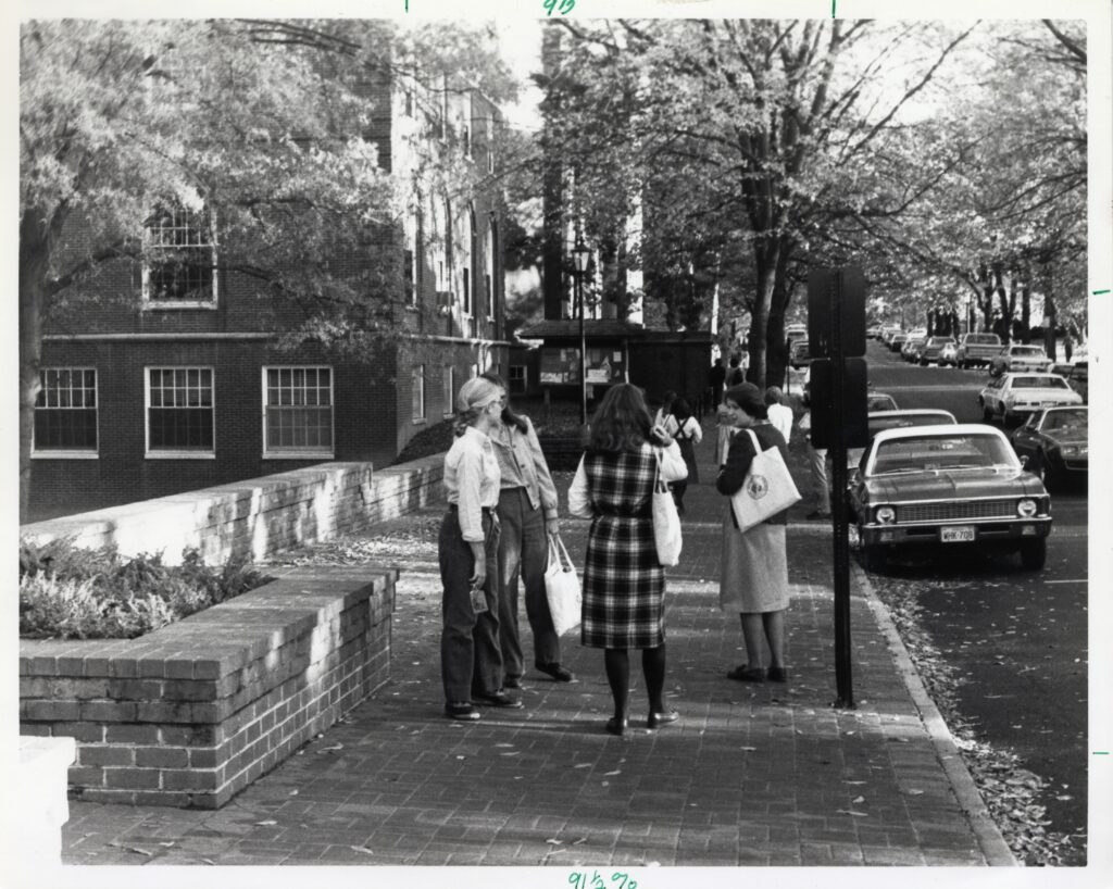 A black and white photo showing a group of students standing on the sidewalk with Campus Drive to the right with cars driving and parked on it. 