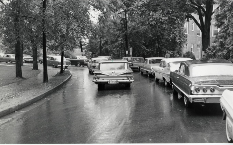 A black and white photo of cars driving on Campus Drive with cars parked on the right side and a sidewalk lined with medium sized trees on the left side. 