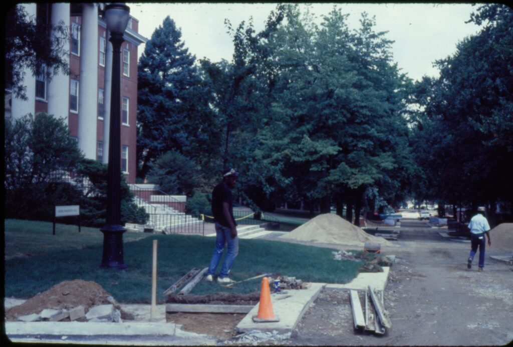 A color photo of the construction work transforming Campus Drive into Campus Walk. Two construction workers are pictured and path has no bricks nor asphalt, but piles of dirt and asphalt on the sides of it. 