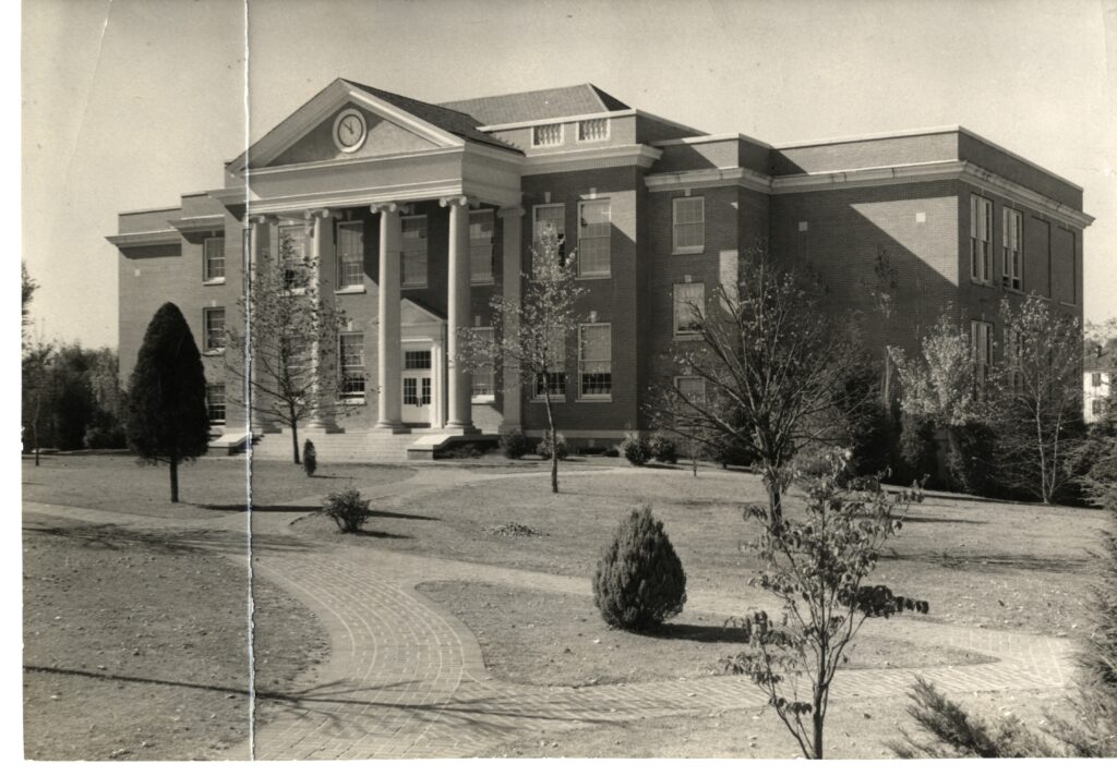 A black and white picture of the two floored Chandler Hall showing its four columned temple entrance and the brick paths in front of it.