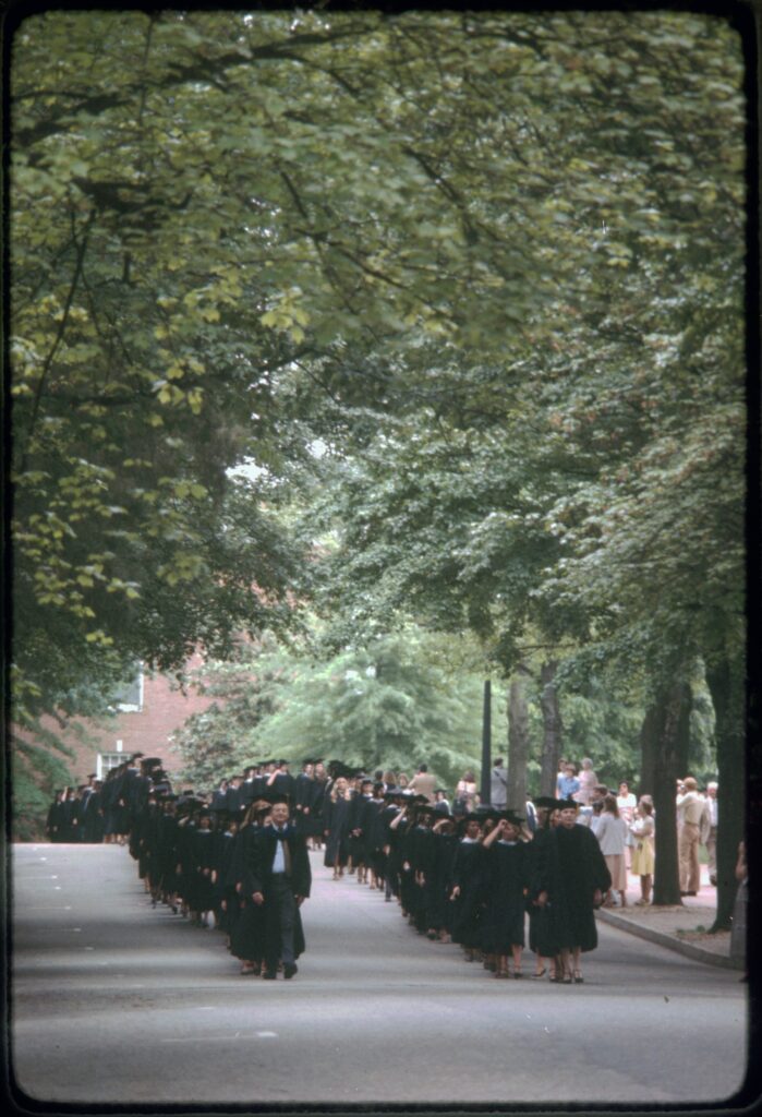 A color picture of students in caps and gowns walking down campus drive in two lines with people taking photos and onlooking on the sidewalk on the right side. 