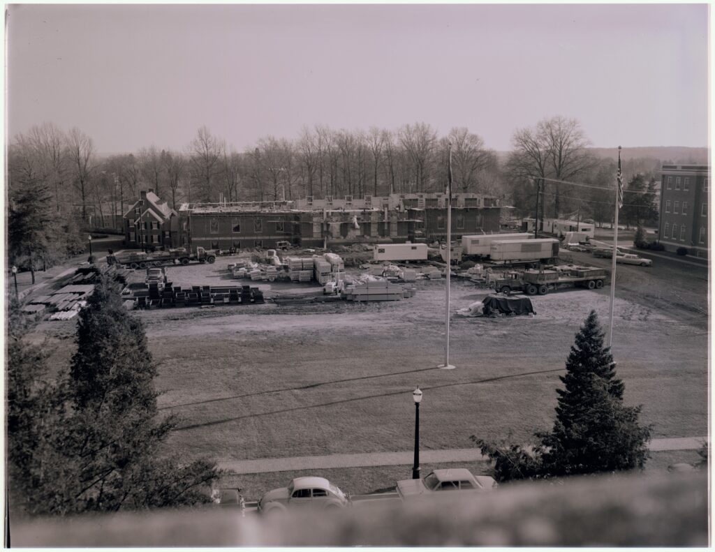 A black and white photo overlooking Double Drive and Jefferson Square with two flagpoles. Behind the two flagpoles, Jefferson Hall is under construction with only the first floor being established at this point and three or four trailers in front of it with building materials scattered around and Framar house in the background.