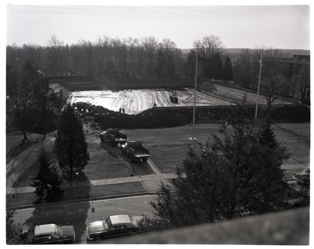 A black and white photo overlooking Double Drive with a line of parked cars on it and Jefferson Square with the rectangular city reservoir taking most of the space of it. There are three pick up trucks on roads from Double Drive towards the reservoir and the bushes surrounding the reservoir have been torn down to make room for workers to deconstruct the reservoir. The reservoir is empty and has a tarping laid out where the water once was. There are two flagpoles in front of the reservoir and Combs Hall stands to the right of it. 