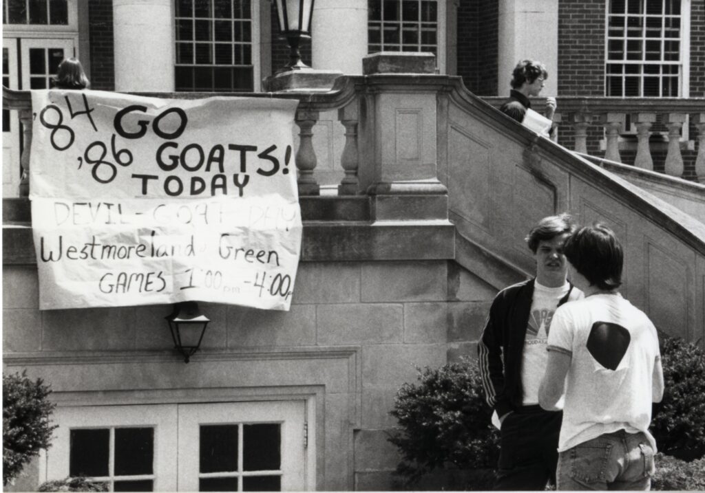 A black and white photo showing the entrance to the Underground in front of the Lee Hall staircase with the two curved stairs on either side. There is a sign that is hanging over top the door to the underground that says "'84 & '86 Go Goats! Today Devil-Goat Day Westmorland Green, Games 1:00 - 4:00."