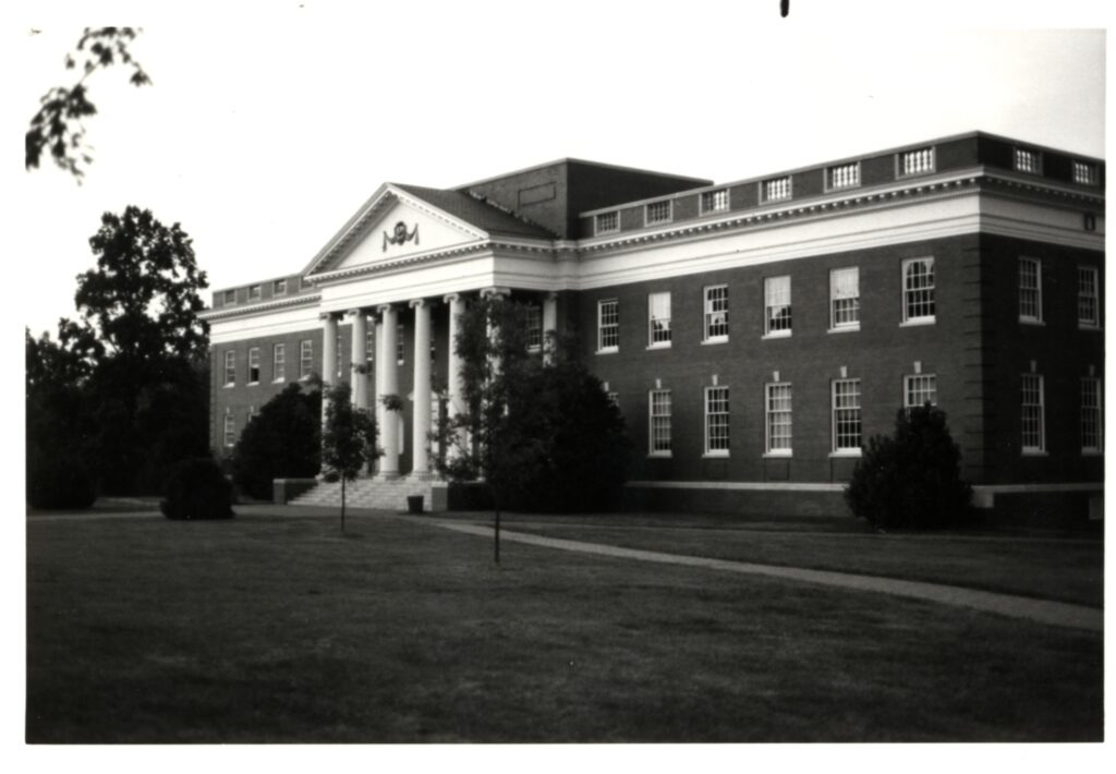 A black and white photo of Dupont Hall showing the still young and small trees and boxwood bushes out front of it.