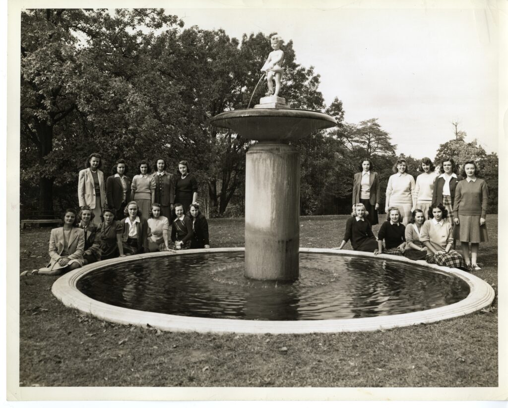 A black and white photo of a circular fountain with a pedestal at the center with an enlarged dish showing a cherub pouring water into it. On either side of the fountain behind it, are several female students. 