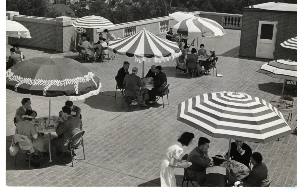 A black and white photo showing many people sitting at four person round tables with patterned umbrellas from the middle of the tables shading them. They are on top of the roof of George Washington Hall.