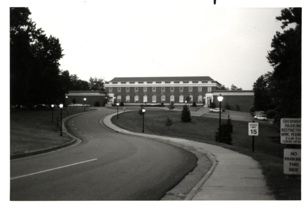 A black and white picture of Goolrick Gymnasium taken from College Avenue. It shows the curvy driveway up to the gym with lampposts and a 15 MPH Speed limit sign.