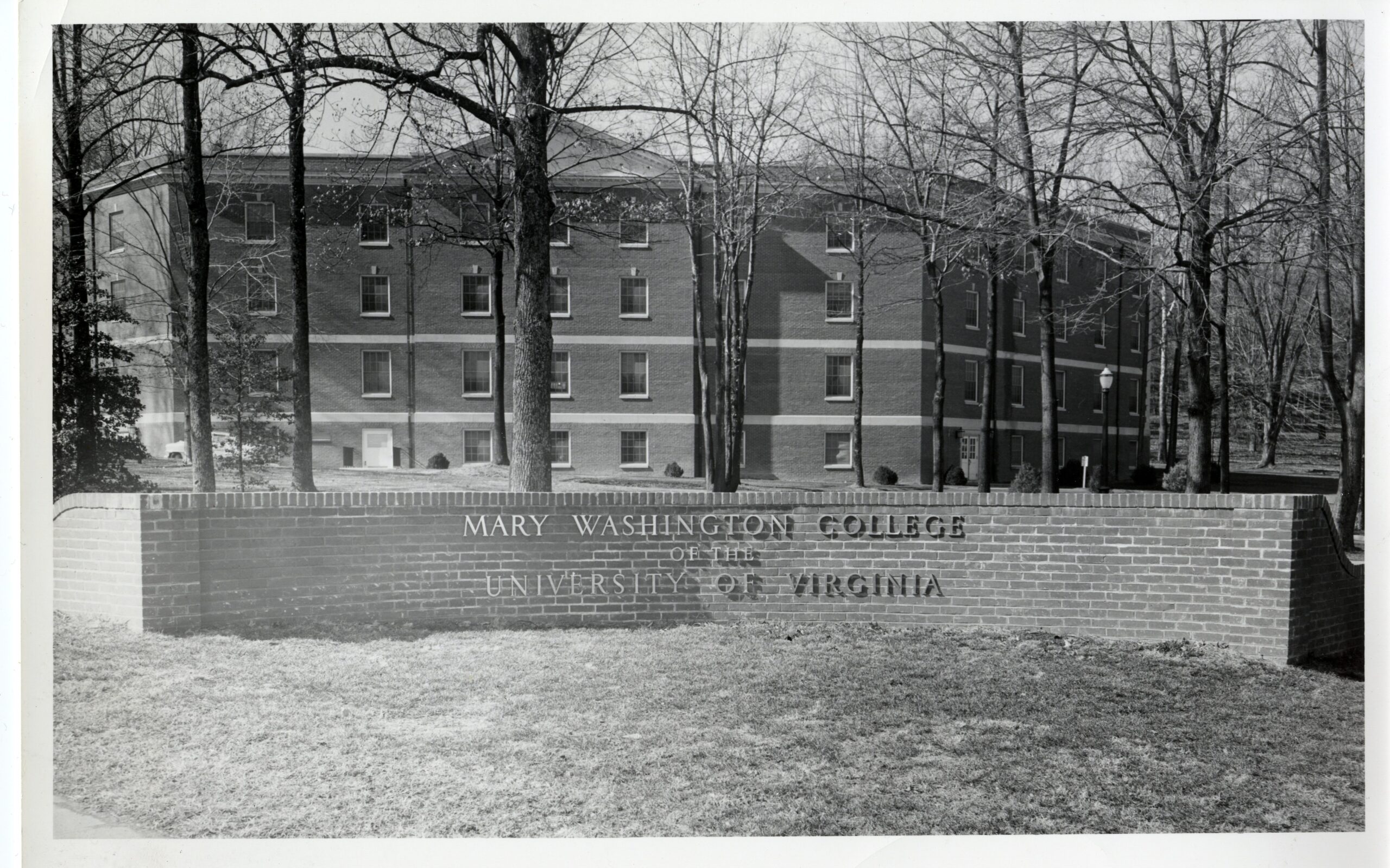 A black and white photo of a curved brick sign Marshall hall standing in the background. The sign reads "Mary Washington College of the University of Virginia."