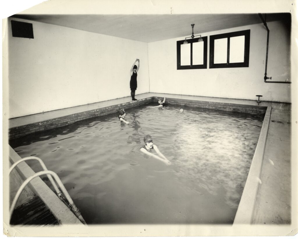 A black and white picture of an indoor swimming pool located within Monroe Hall. Four women are swimming in it with one getting ready to dive in.