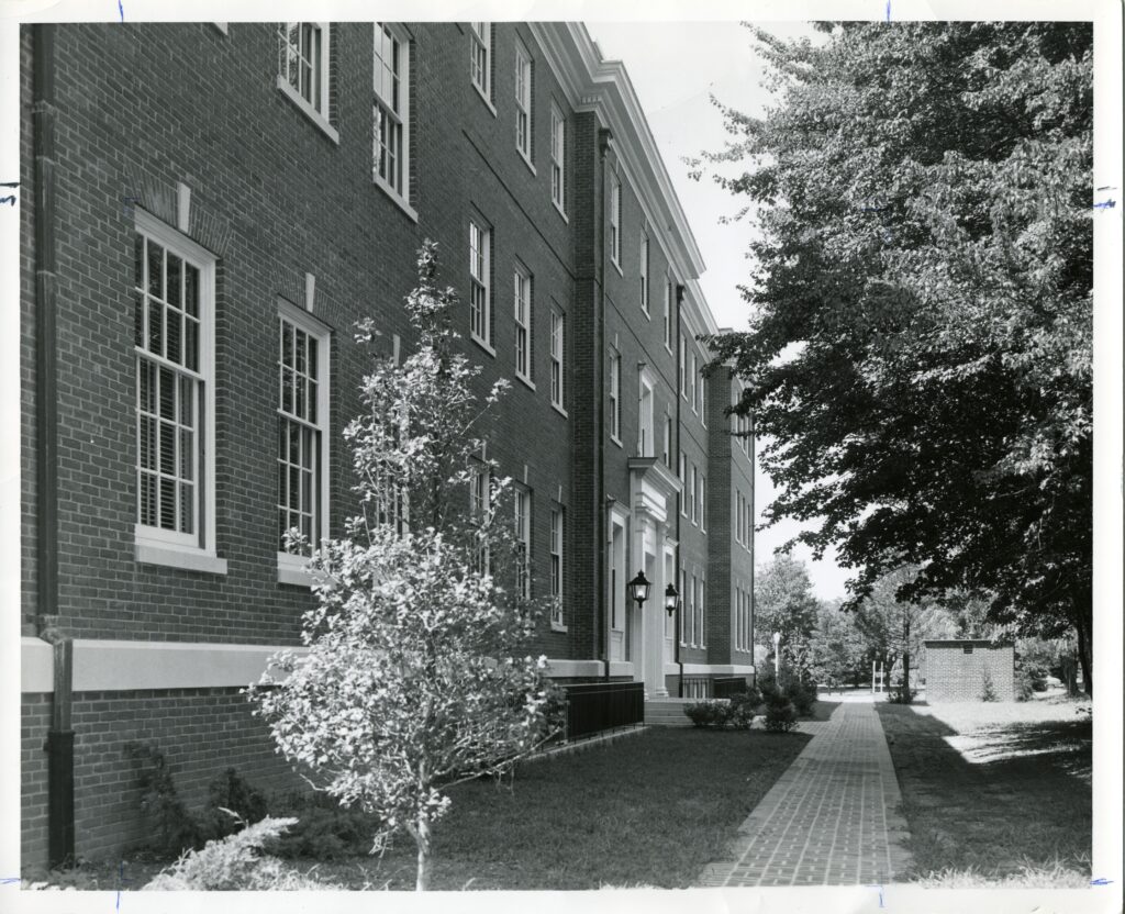 A black and white photo showing a brick pathway with Combs Hall on the left. There are small trees on the left side of the pathway and larger trees on the right side with a small brick structure in the distance. 