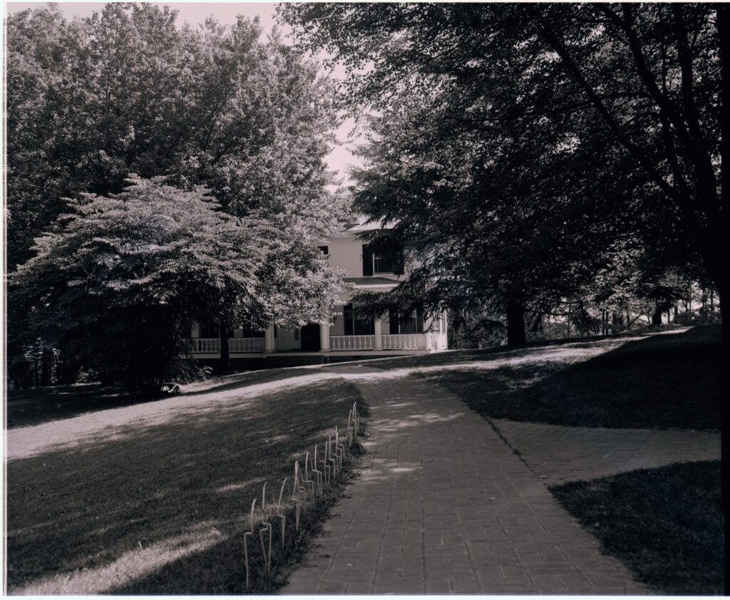 A black and white photo of a brick pathway leading to Marye House. There are large trees shading the path and Marye House has a covered porch with a white fence and white columns.