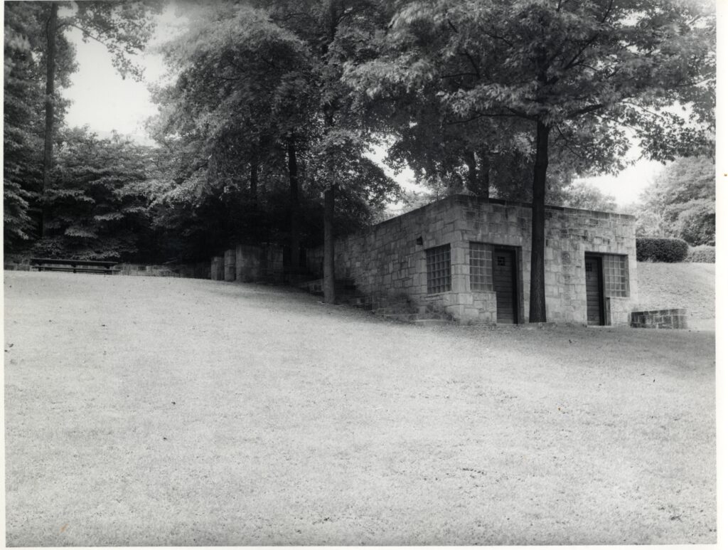 A black and white picture taken downhill from a concrete patio on the edge of a woods with a cinderblock and glass block building at the right side of the image. There is freshly cut grass heading up to both of them. 