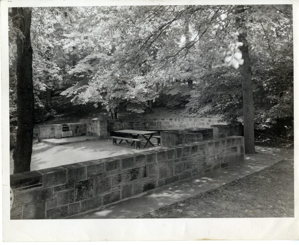A black and white photo showing a concrete patio with a picnic table and grills built into a cinderblock wall. The whole area is shaded by large trees. 