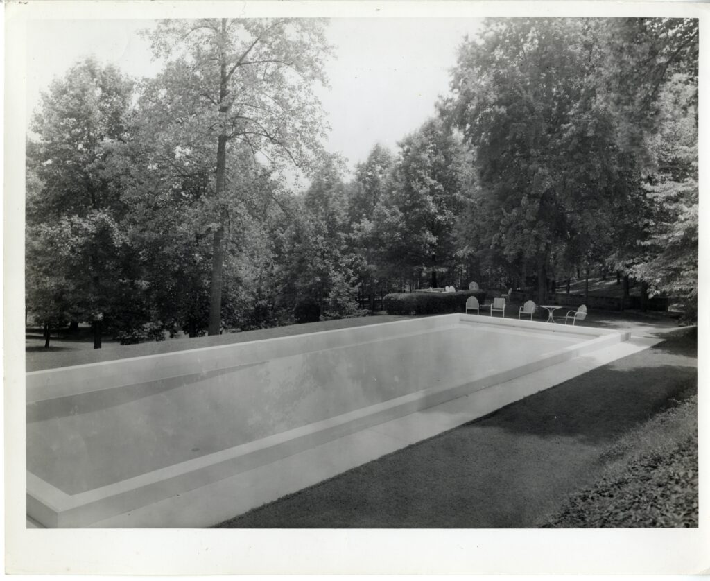 A black and white photo showing a rectangular pool with four pool chairs and a table at the other end of it. The pool is surrounded by grass and larger bushes and trees on the far edges.