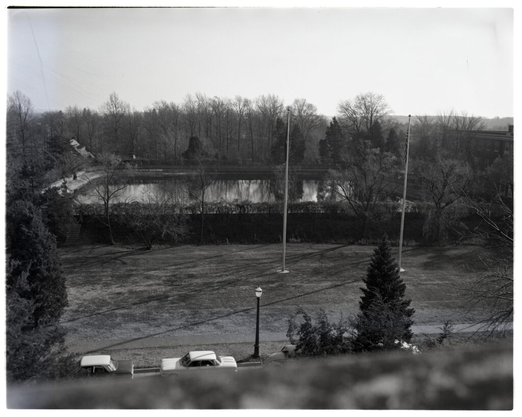 A black and white photo overlooking Double Drive with a line of parked cars and Jefferson Square, complete with two flagpoles and the city reservoir surrounded by bushes and large trees without leaves and Combs Hall to the right. 