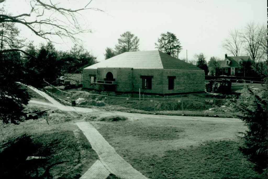 A black and white picture showing a rectangular building with a hipped roof still under construction; it only has cinderblocks but no siding and nothing where the windows should be. The building has a chain-link fence around it and a backhoe on the right side of the building. There are brick paths leading up to the building and around it. 