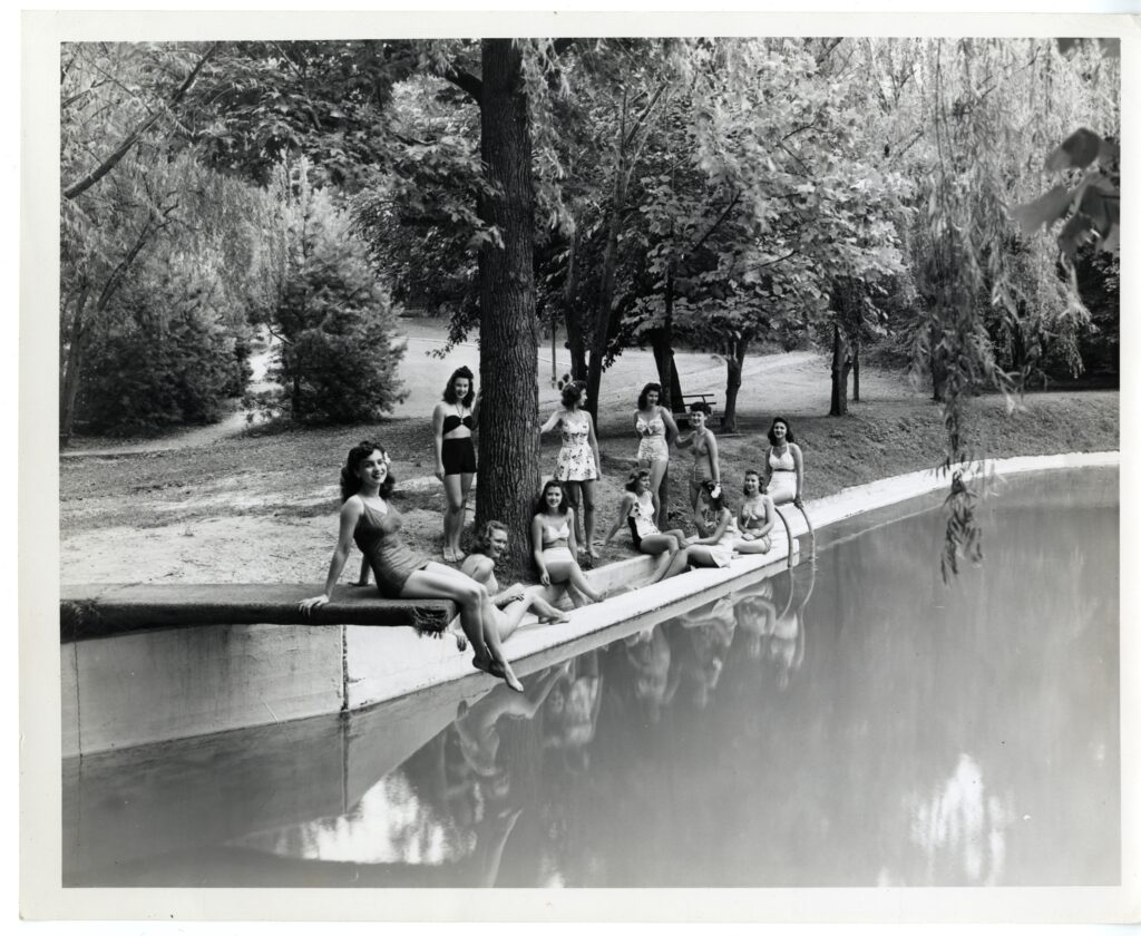 A black and white picture of 11 female students relaxing on the edge of the outdoor swimming pool underneath some shaded trees. The pool has curvilinear edges and there is one student sitting on the diving board.