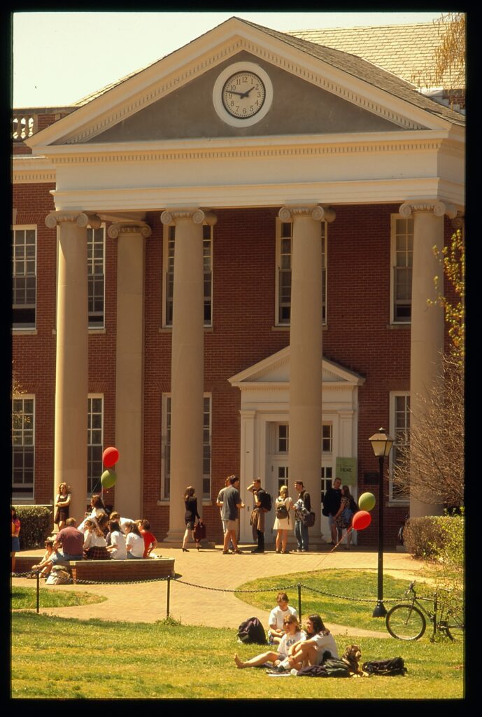 A color photo showing the front of Chandler Hall with many students hanging around out front of it and a few balloons around them.