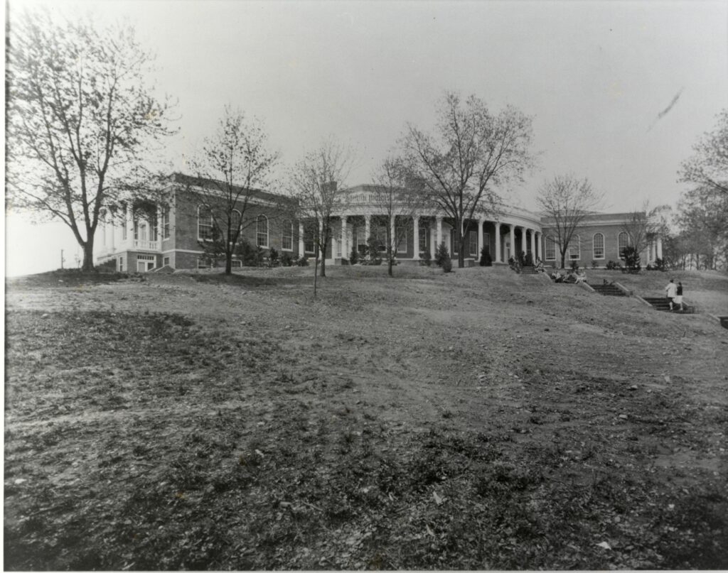 A black and white photo of Seacobeck Hall taken from the bottom of the grassy hill. There are students walking up the stairs to the front entrance of Seacobeck Hall.