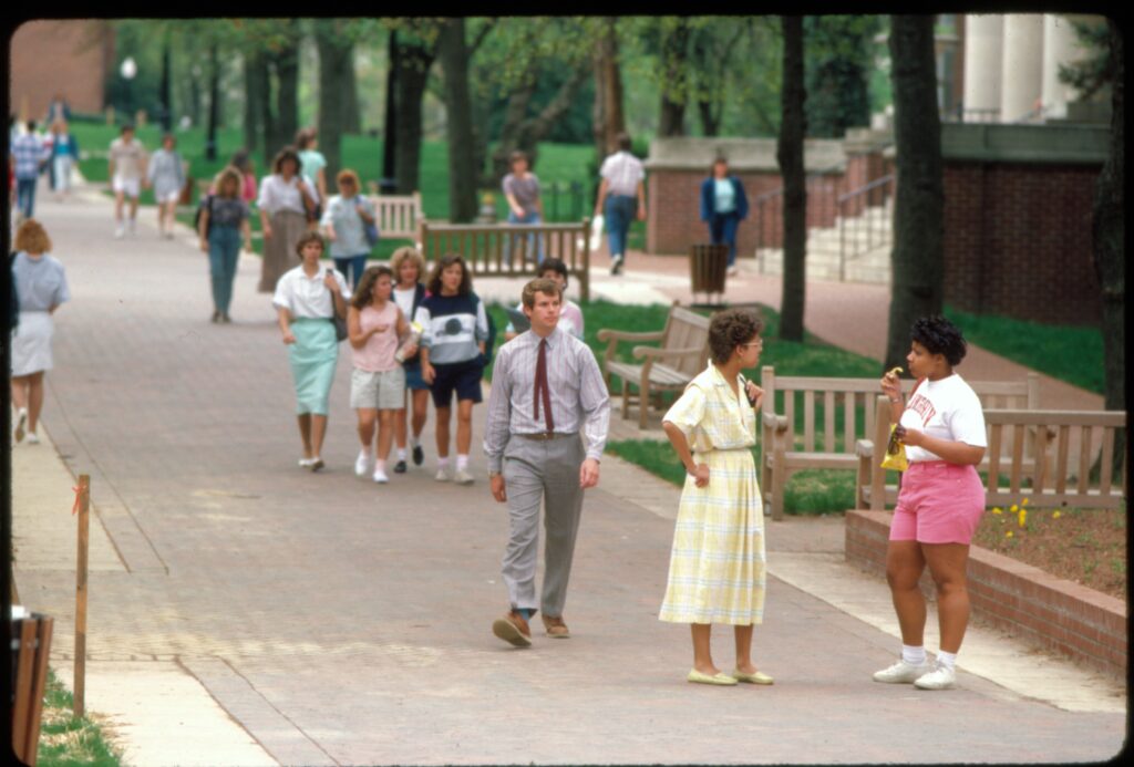 A color photo of students walking up and down the brick Campus Walk with benches along the side and brick flower beds. 