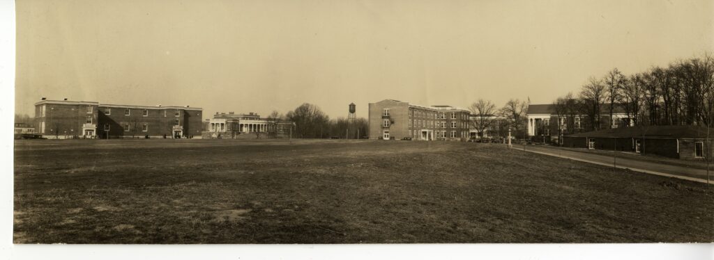 A black and white photo of campus taken from ground level showing the unfinished Chandler and Virginia Halls (both without their finished columned entrance) with the plain alumni hall to the right side with Monroe Hall behind it. In between Virginia and Chandler Halls in the background is Seacobeck Hall and a water tower with trees on the horizon.