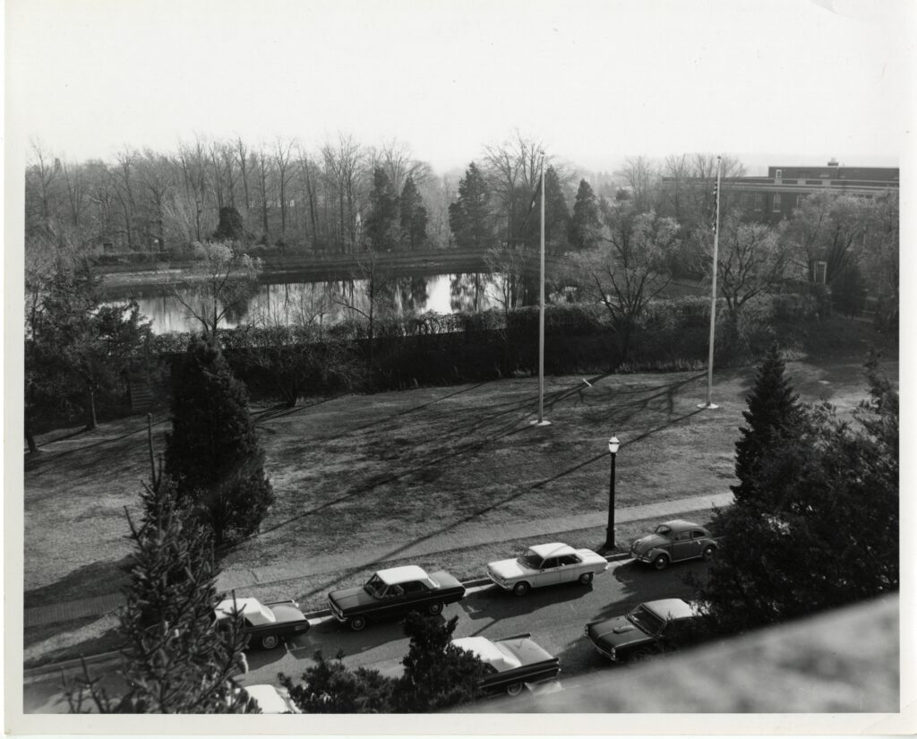 A black and white picture overlooking double drive with  cars parked on both sides and showing the city reservoir standing at what is now present day Jefferson Square with the two flagpoles in front of it. The reservoir is surrounded by greenery and Combs Hall stands to the right of the reservoir
